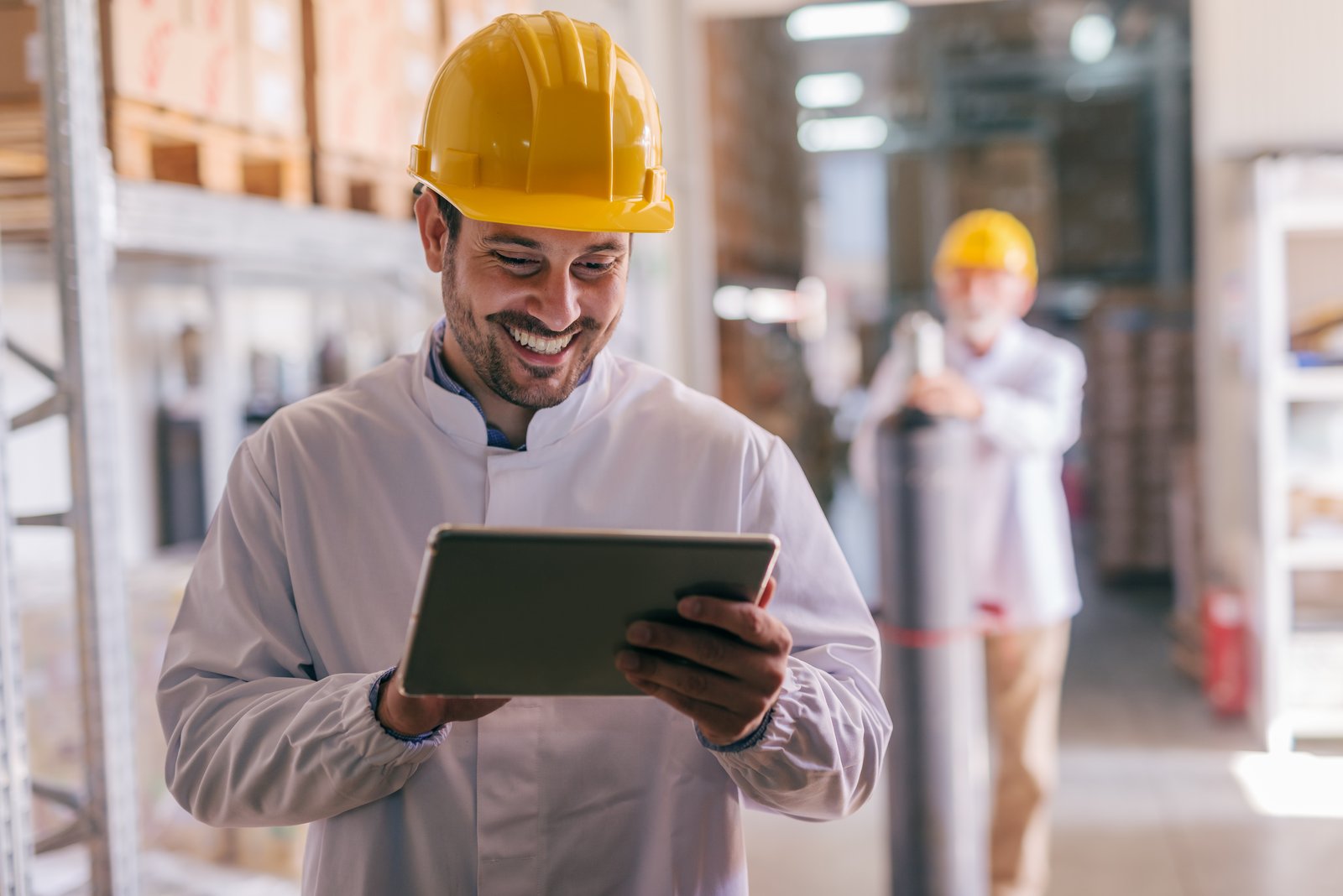 Man looking at a tablet laughing and wearing a construction site helmet in a warehouse. In the background another man