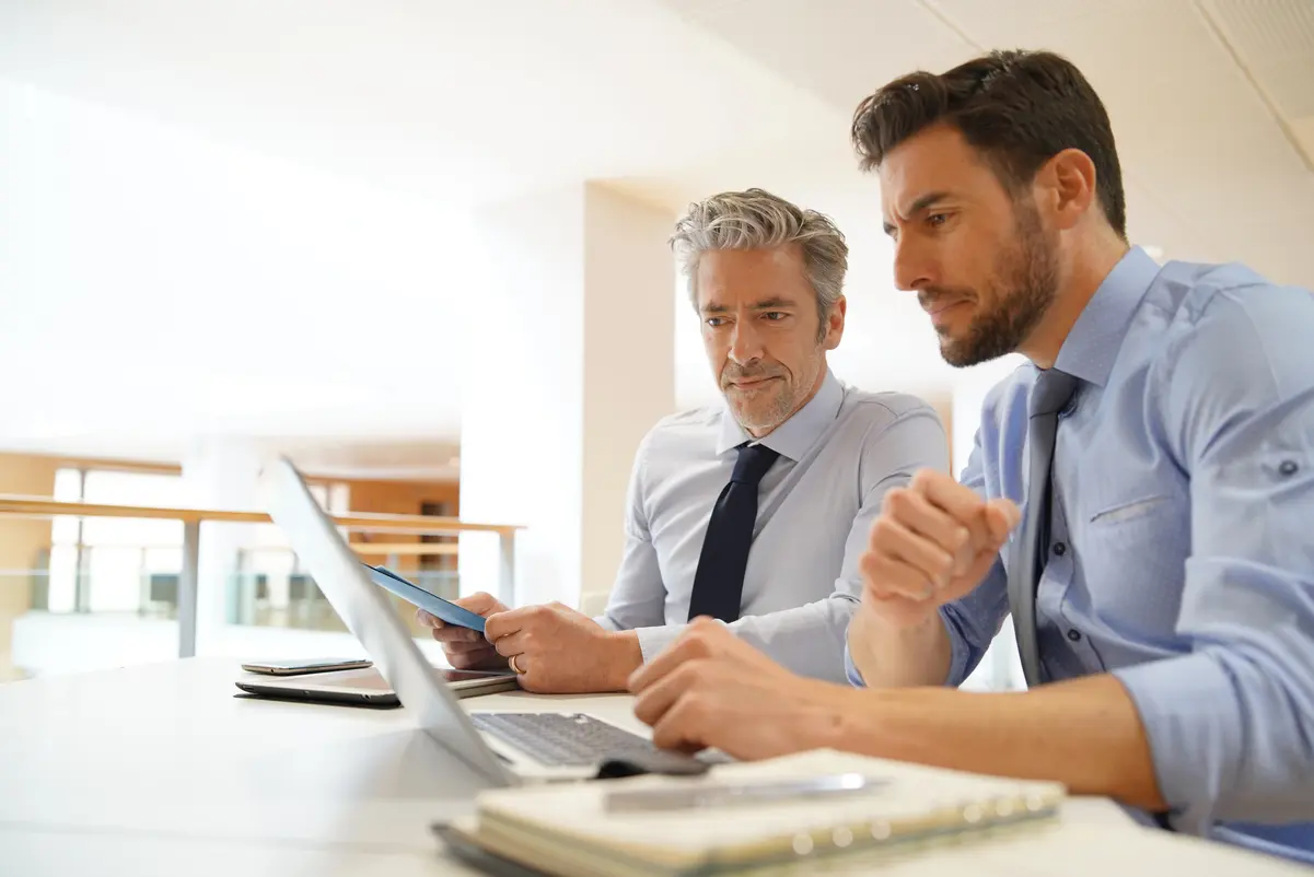 2 men at a desk looking at a laptop, one man pointing at the screen