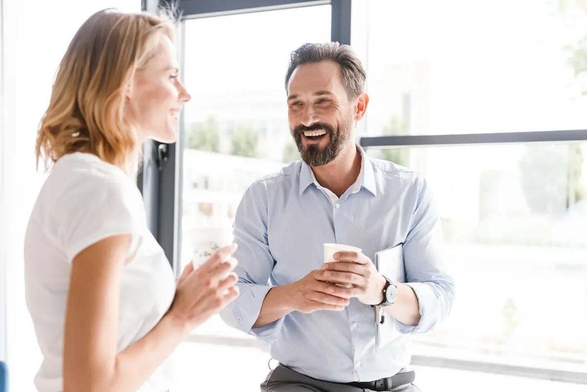 Man and woman standing in an office laughing, in the background a glass front in sunshine