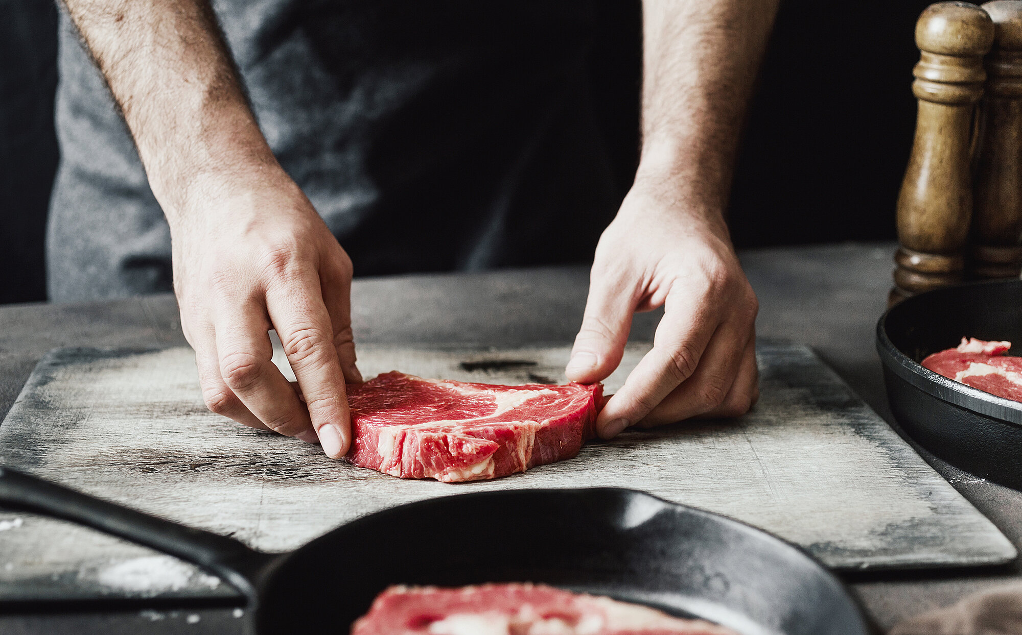 Man processing a piece of meat, in the foreground a pan, on the right a pepper shaker