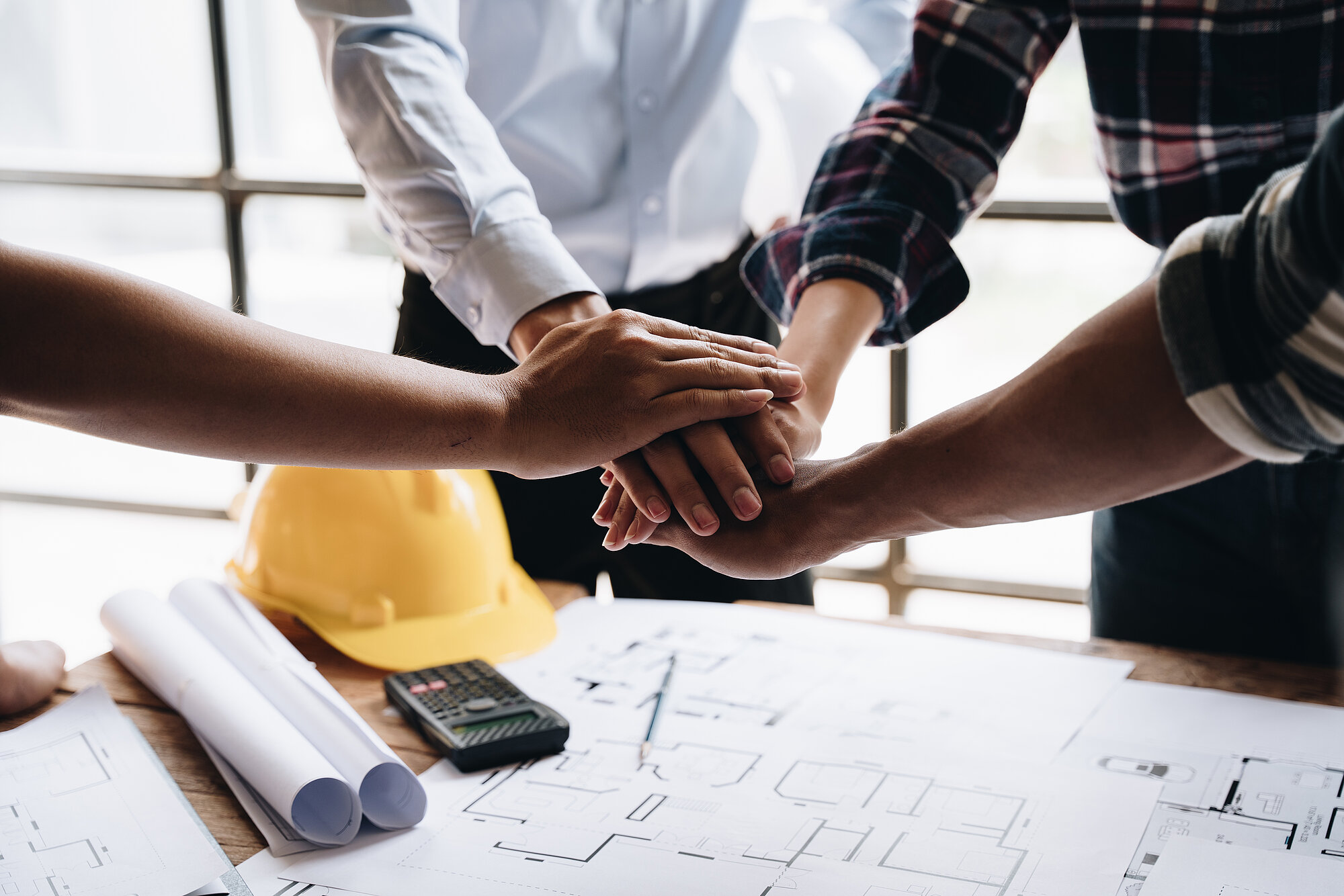 People lay their hands on each other, along with a table with building materials and the utensils of a building contractor such as plans, calculators and pens