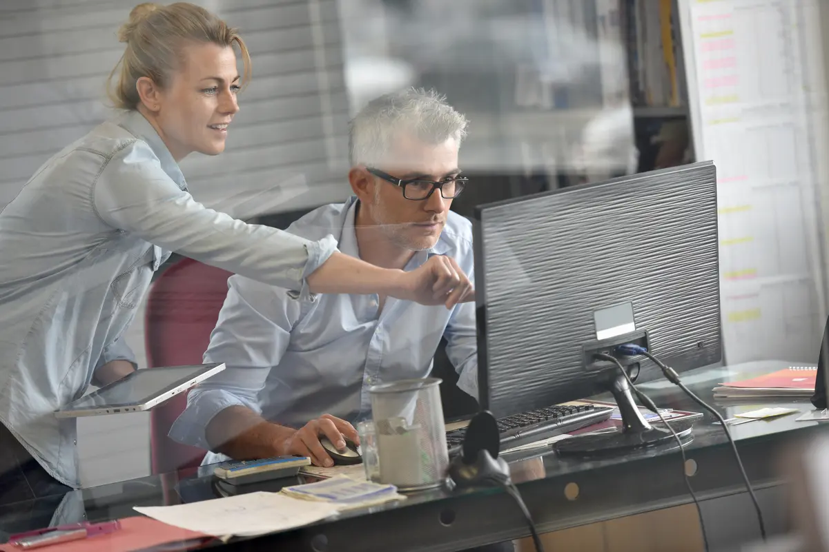 Man and woman in an office , where the woman is standing and pointing at the screen and the man is sitting and looking at the screen