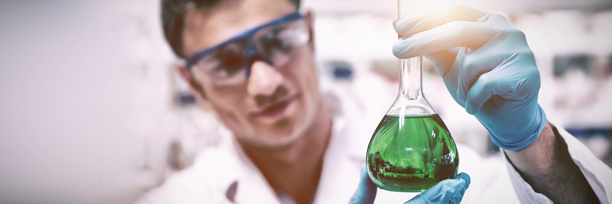 Chemist with safety glasses, and gloves holding a glass filled with a green liquid